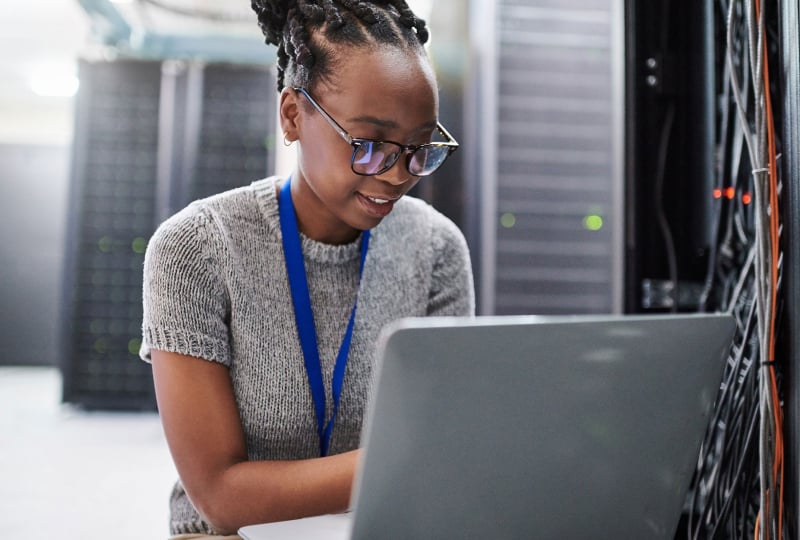 Woman sitting in a server room working on a laptop.