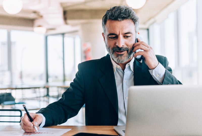 Man sitting at a desk talking on a cellphone and writing down notes with his right hand.